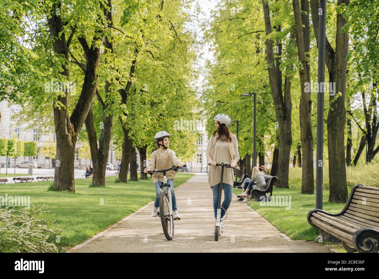 Girl cycling while mother riding electric scooter on road in city park Stock Photo