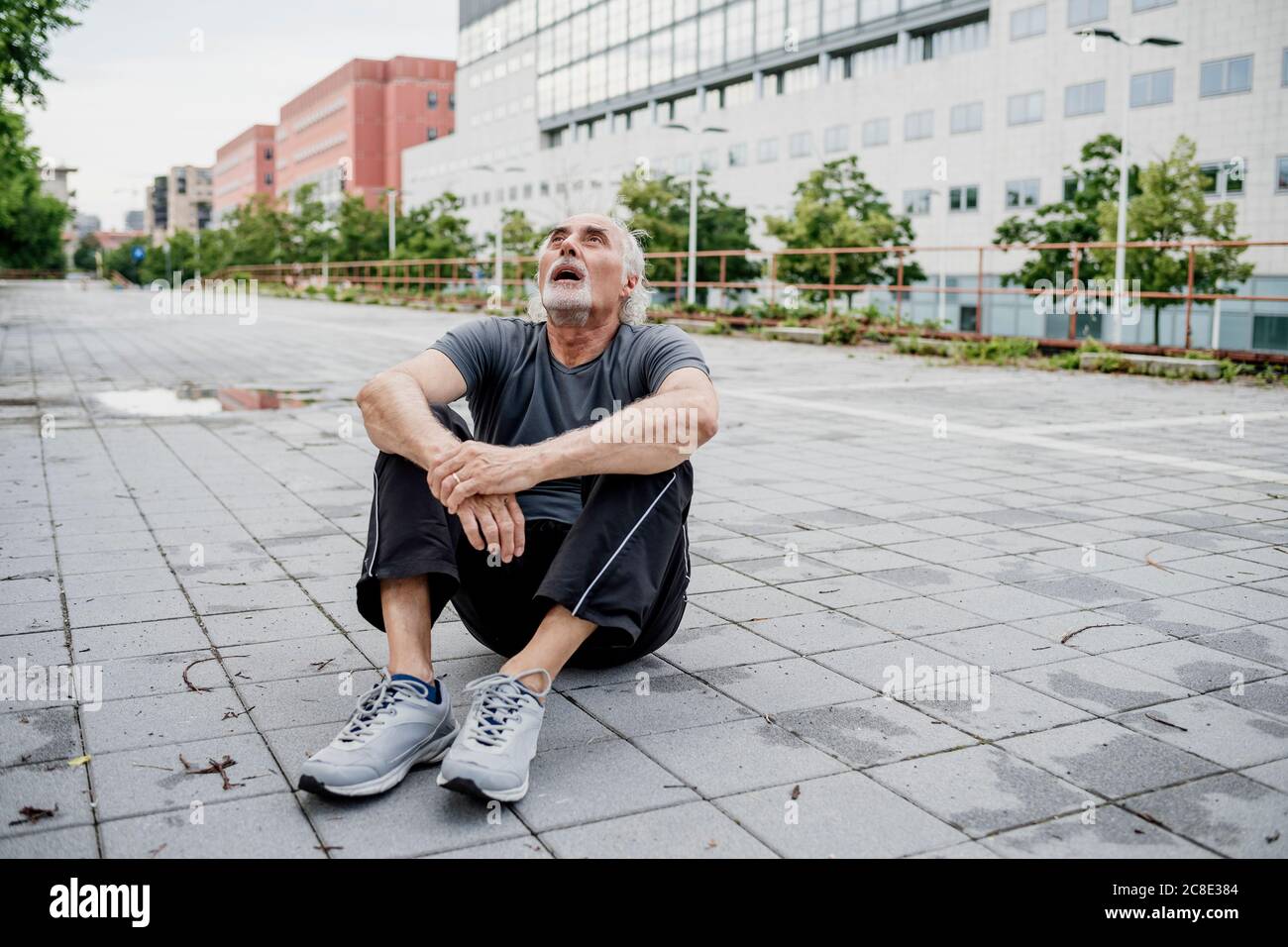 Tired senior man with mouth open looking up while sitting on footpath in city Stock Photo