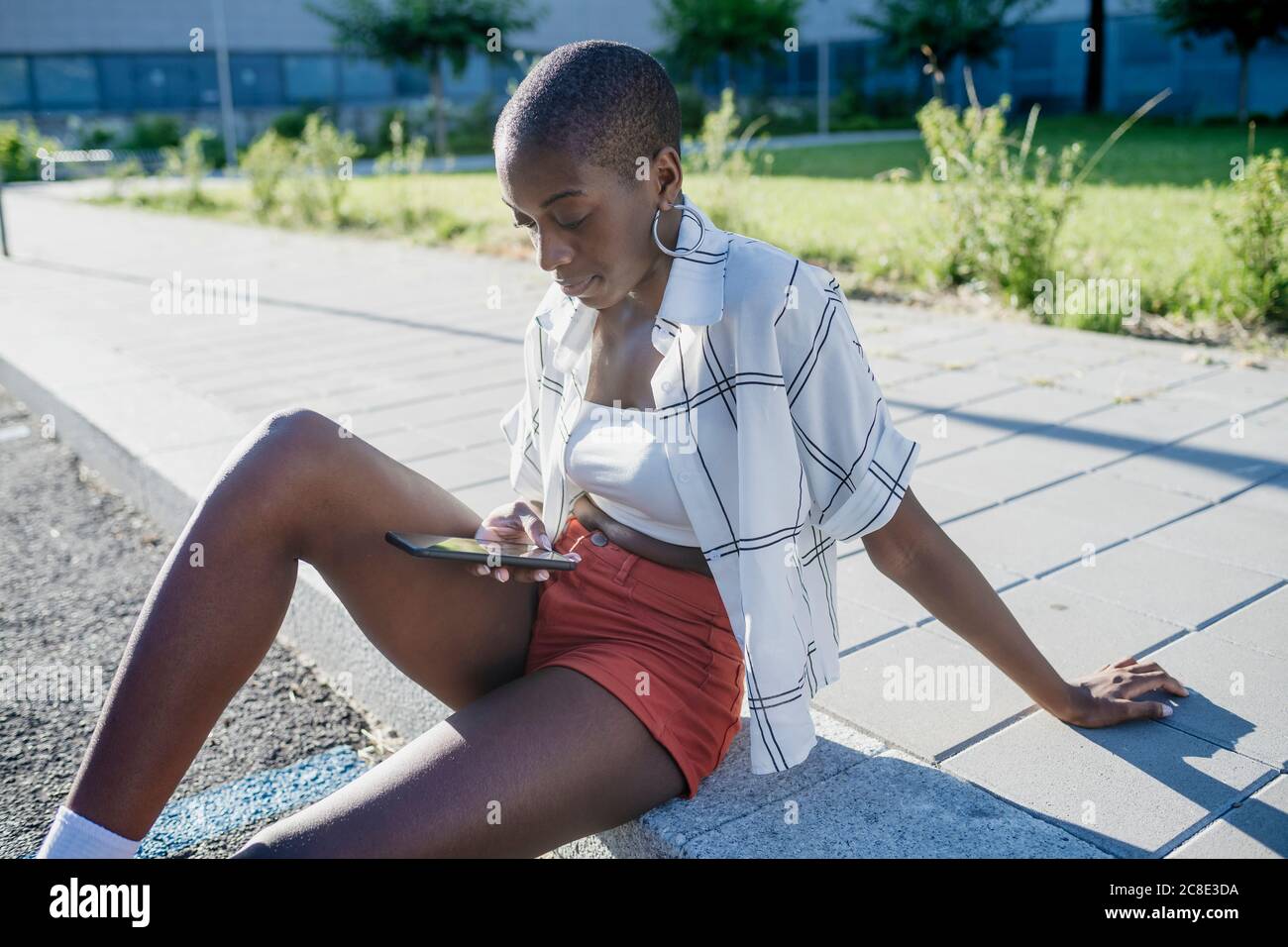 Young woman with shaved head using mobile phone while sitting on footpath in city Stock Photo