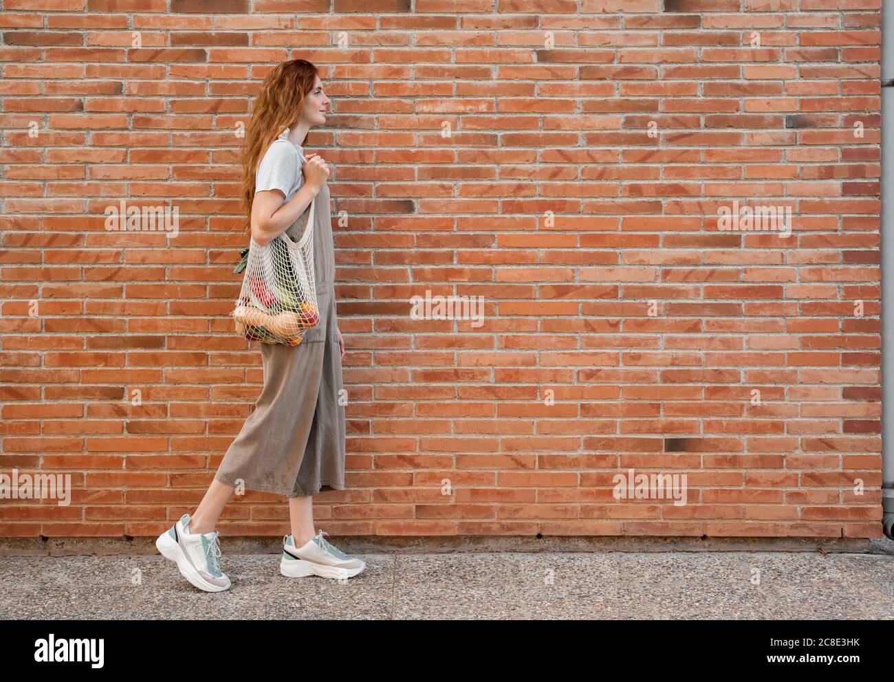 Young woman carrying groceries in mesh bag while walking on footpath by brick wall Stock Photo
