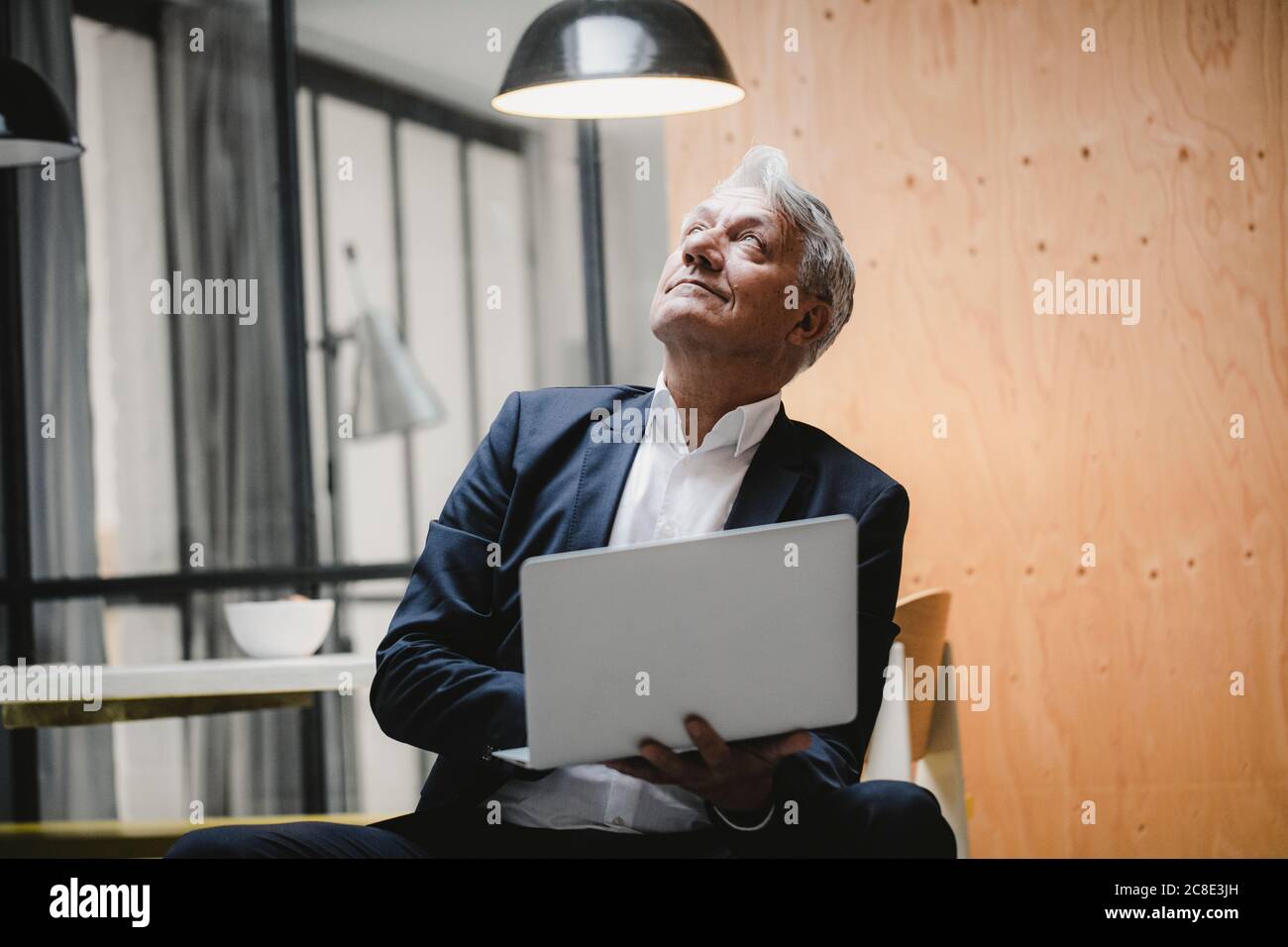 Senior businessman sitting under ceiling lamp, using laptop, looking into the light Stock Photo