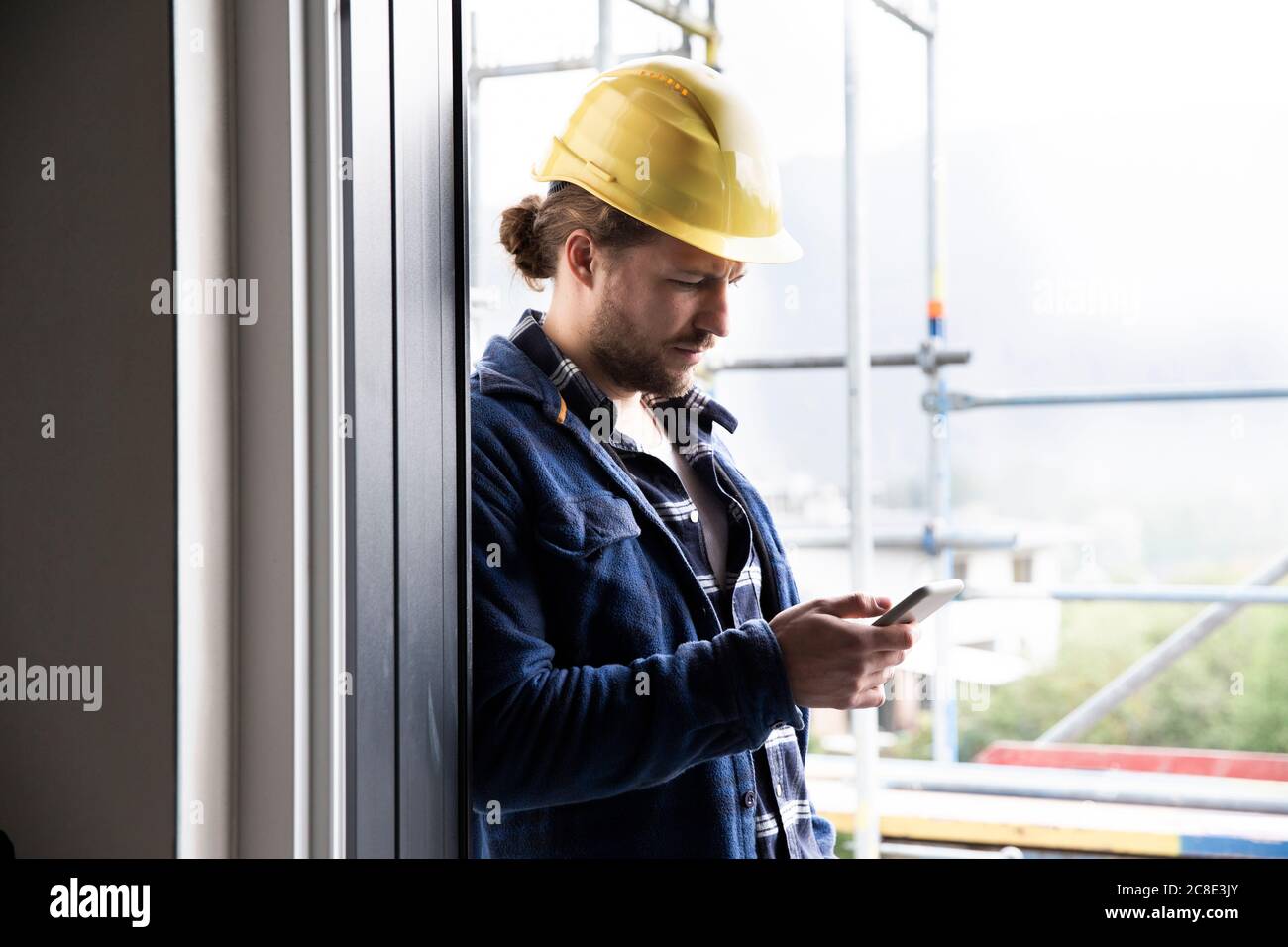 Construction worker wearing helmet using smart phone by window at constructing house Stock Photo