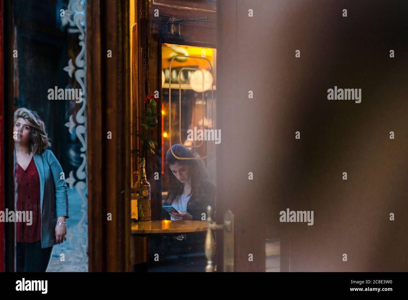 Young woman using smart phone while girlfriend standing at entrance of cafe seen through glass Stock Photo
