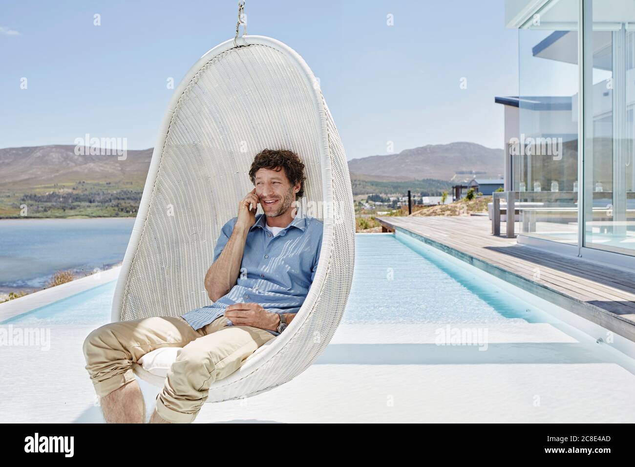 Man sitting in hanging chair above swimming pool talking on the phone Stock Photo