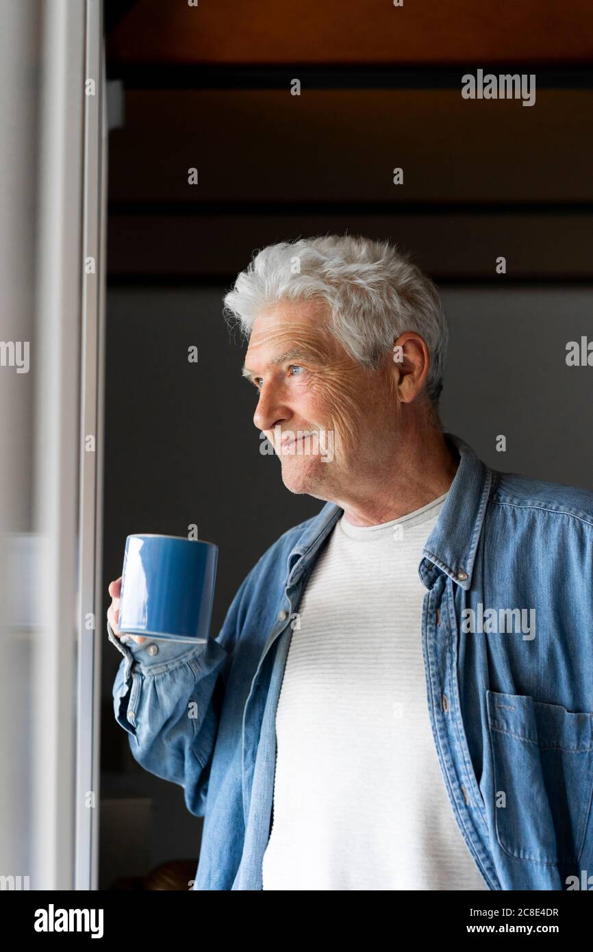 Thoughtful senior man smiling while drinking coffee by window at home Stock Photo