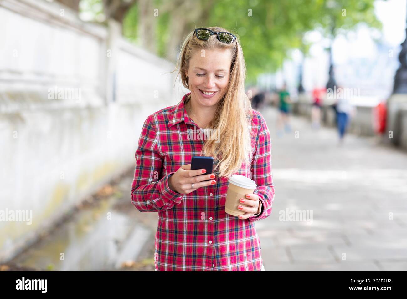 Smiling mid adult woman holding coffee using mobile phone while standing on footpath Stock Photo