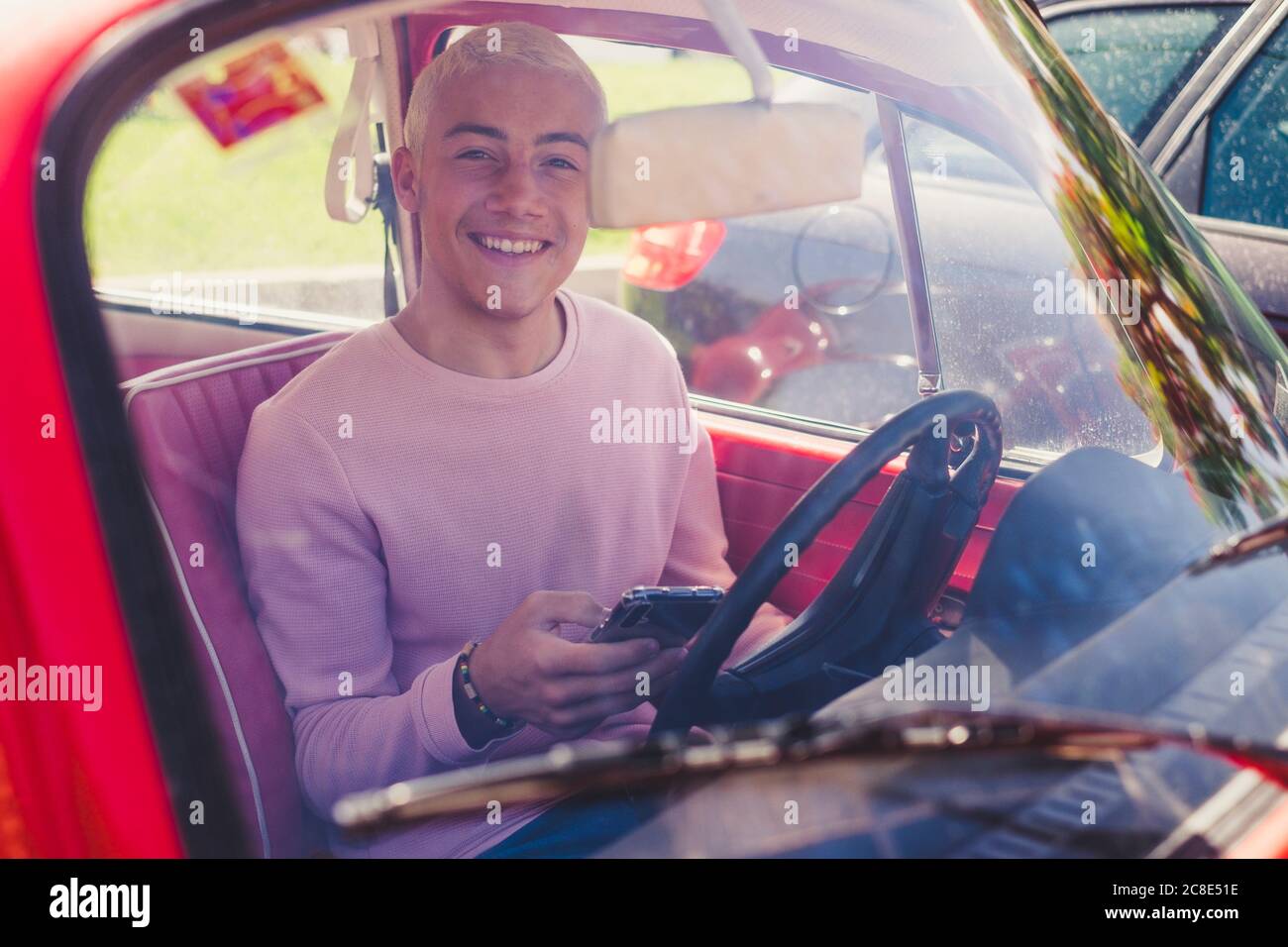 Portrait of smiling teenage boy sitting in vintage car with smartphone looking out of window Stock Photo