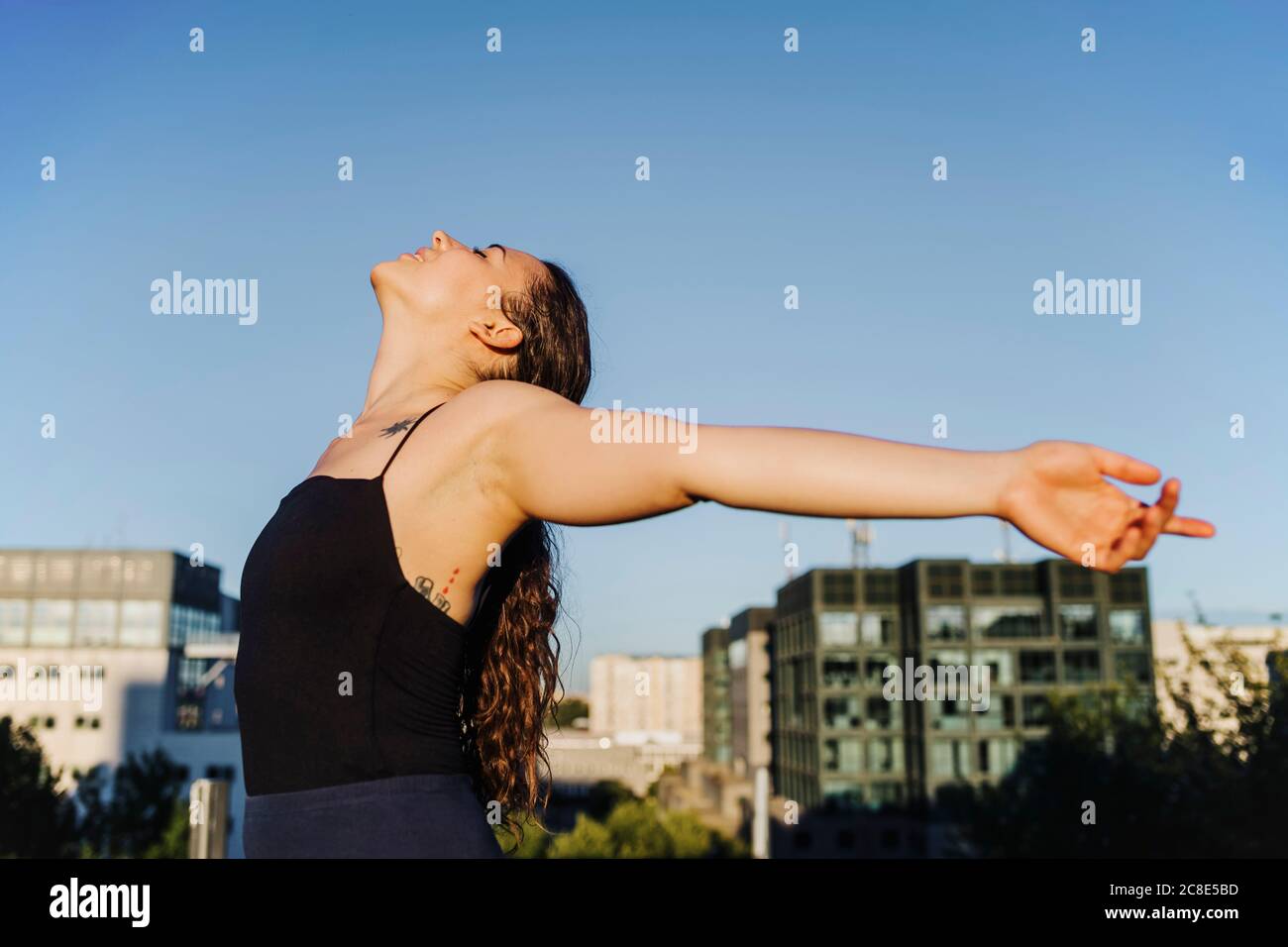 Carefree sporty woman standing with arms outstretched in city against clear blue sky Stock Photo