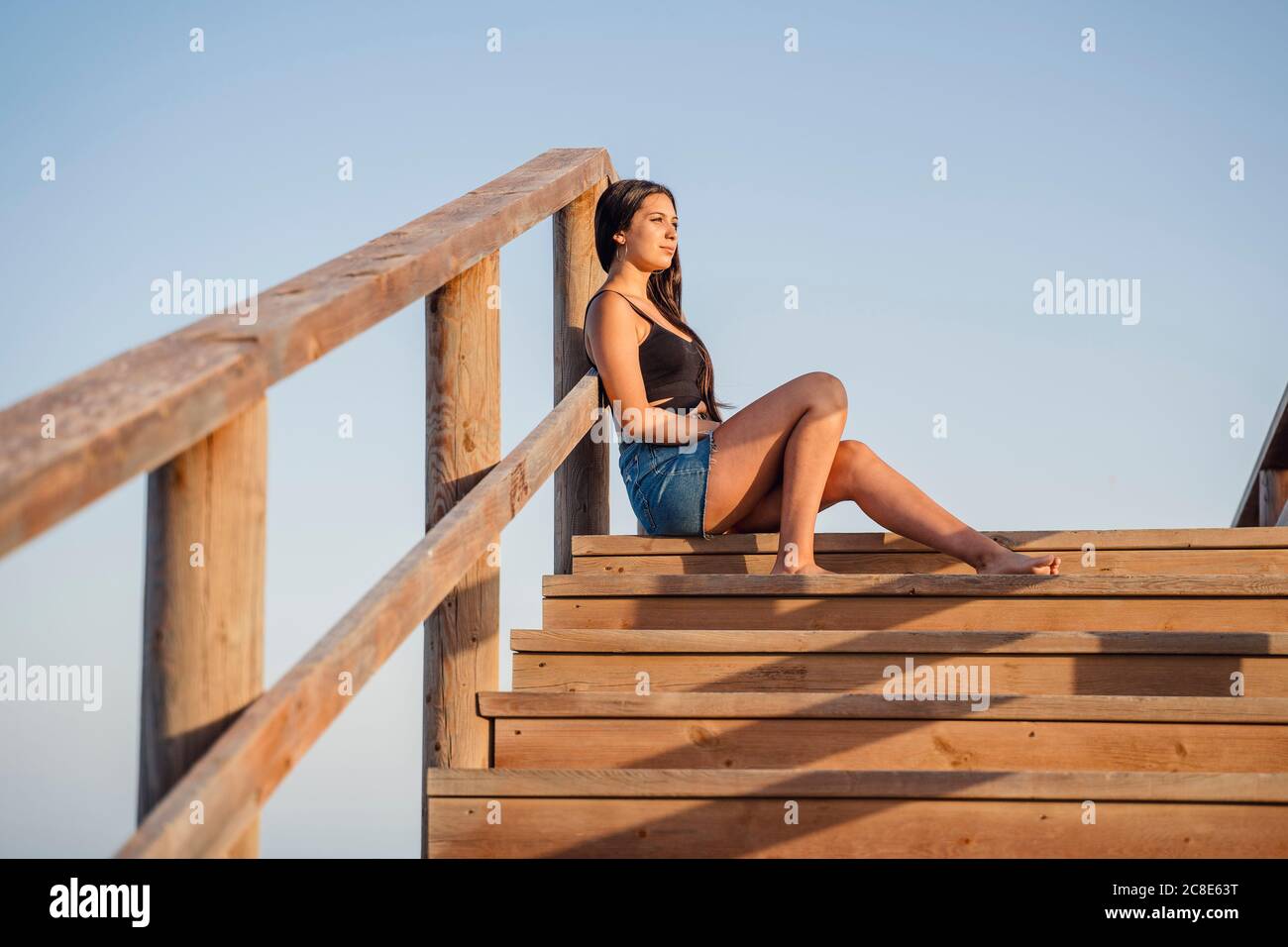 Thoughtful teenage girl sitting on wooden steps against clear blue sky Stock Photo