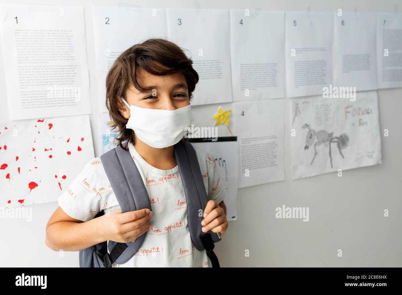 Boy wearing mask standing against papers stuck on wall in school Stock Photo