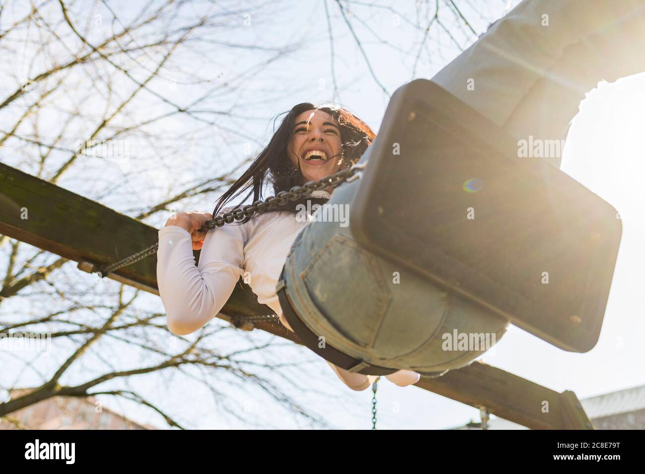 Cheerful young woman swinging against sky in park during sunny day Stock Photo