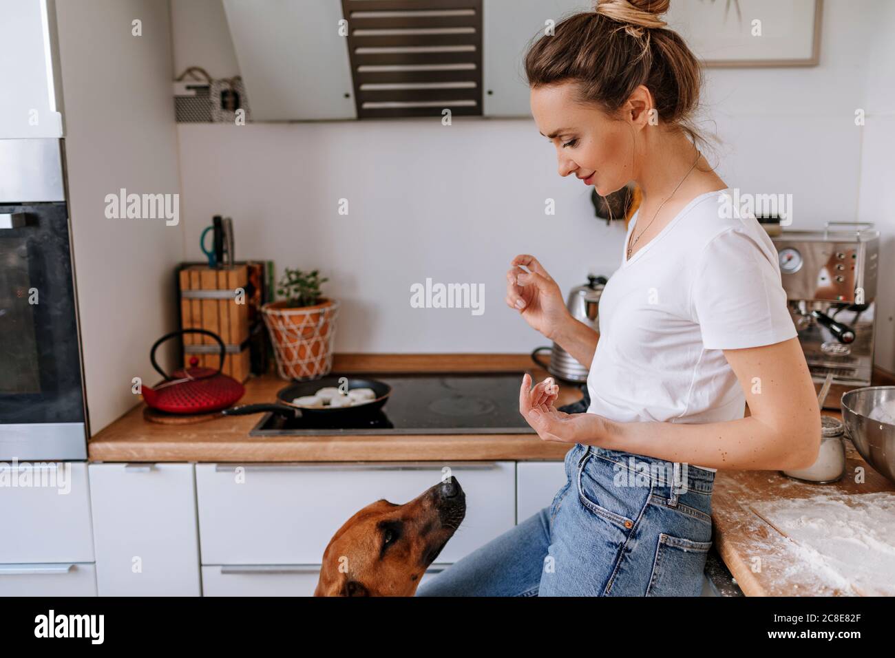 Beautiful woman with messy hands looking at dog while standing in kitchen Stock Photo