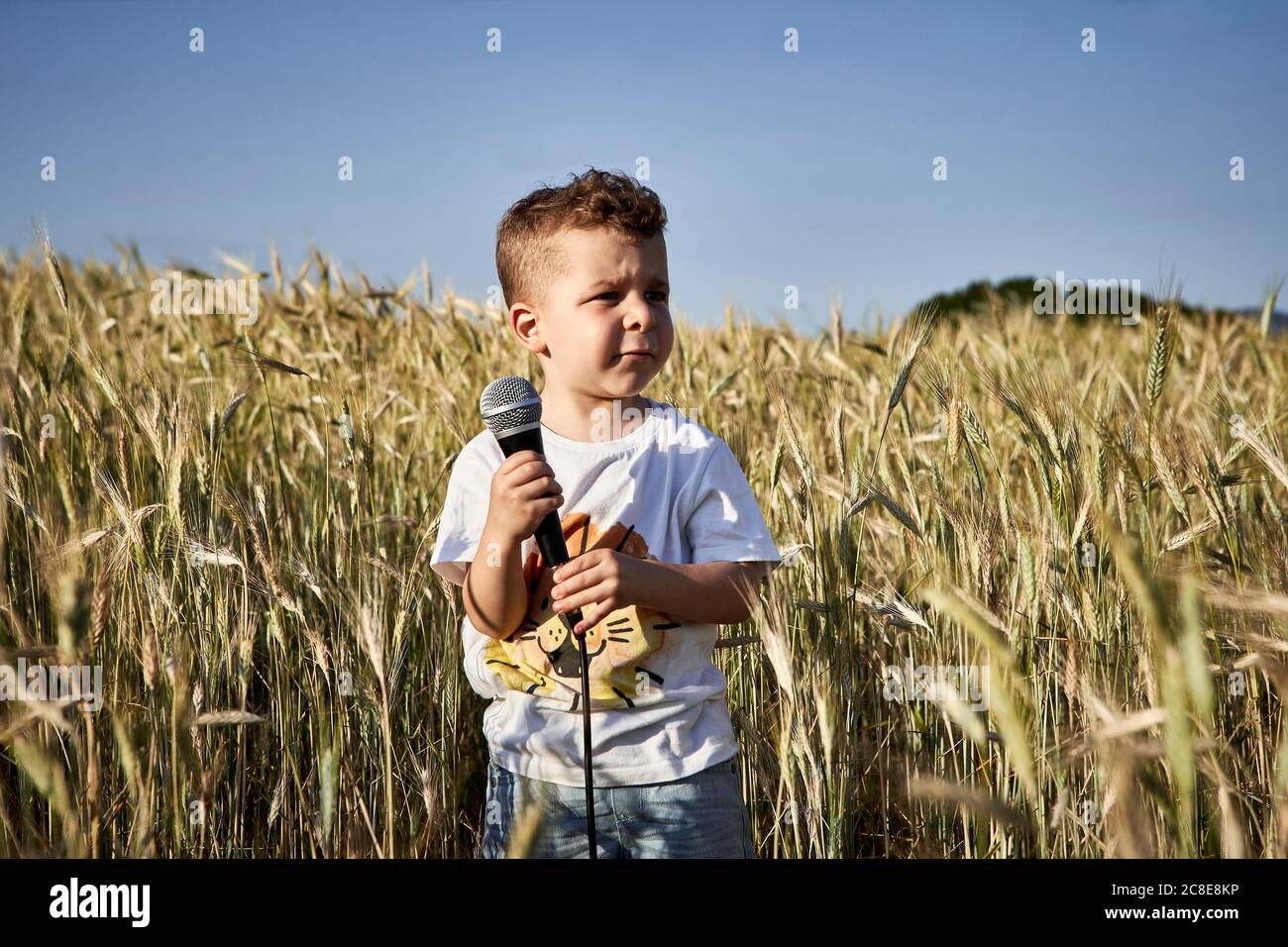 Boy holding microphone looking away while standing amidst crops against clear sky Stock Photo