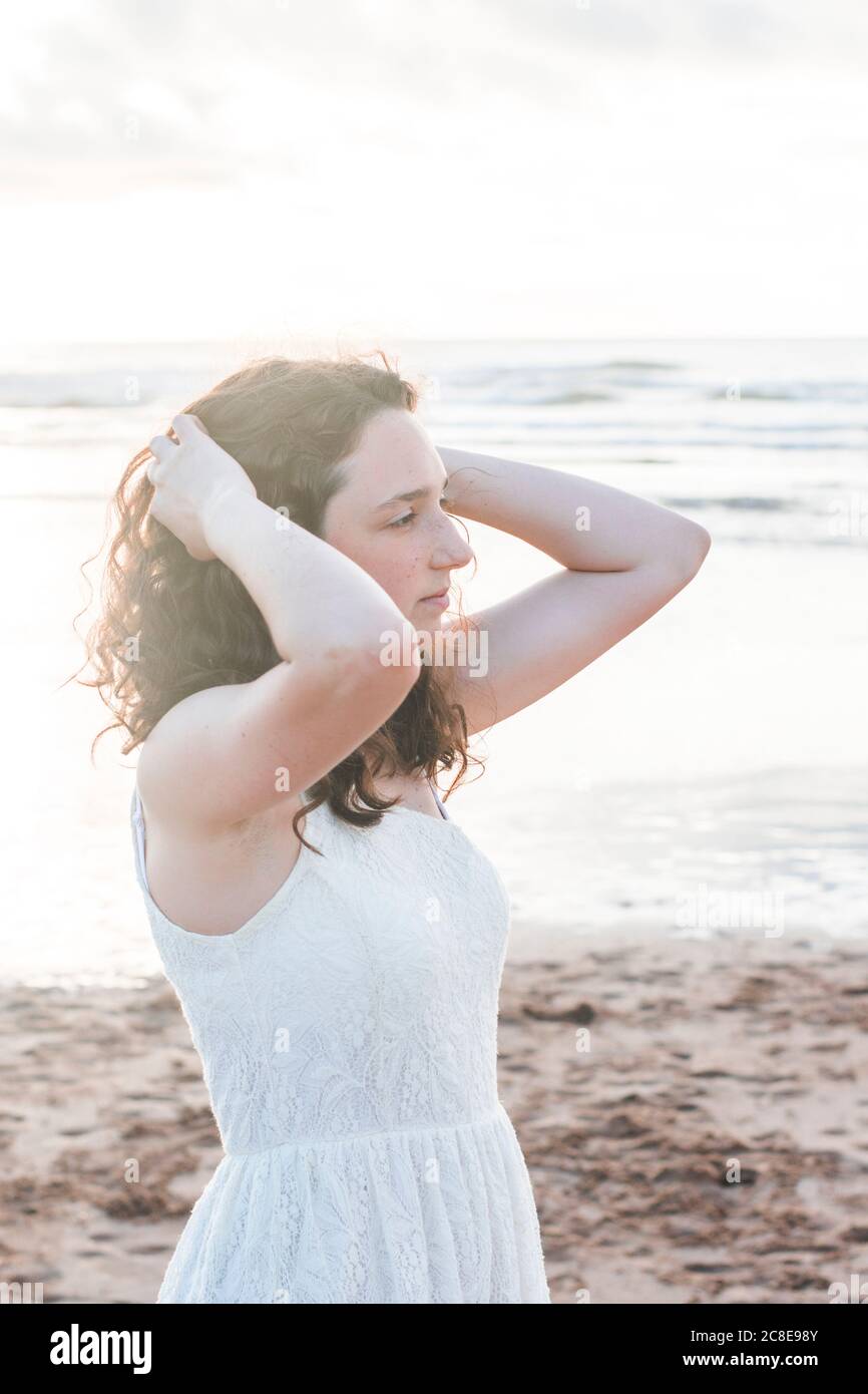 Thoughtful young woman wearing white dress with hands in hair standing at beach Stock Photo