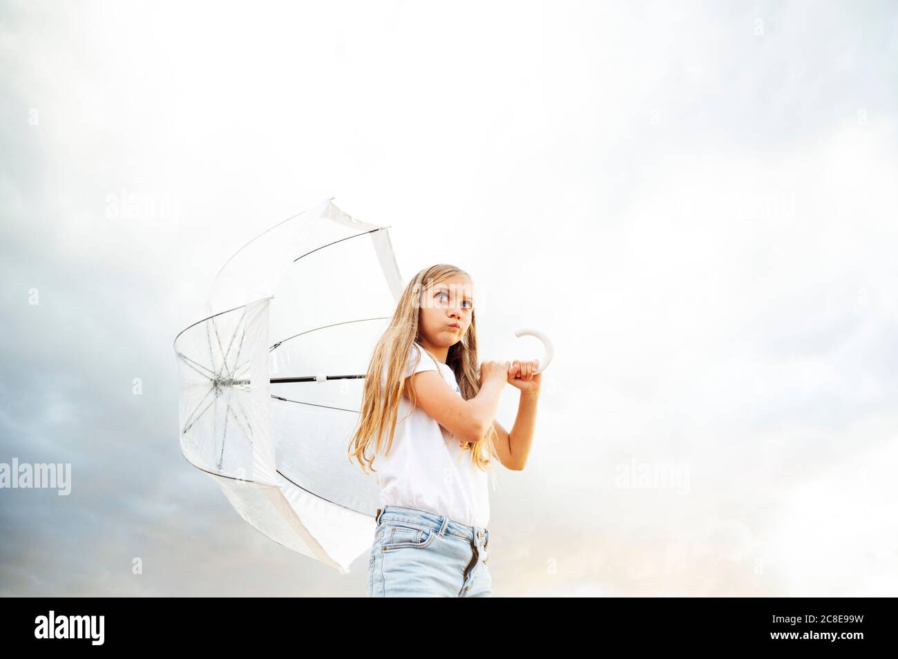 Blond girl making face while holding umbrella and looking away against cloudy sky Stock Photo