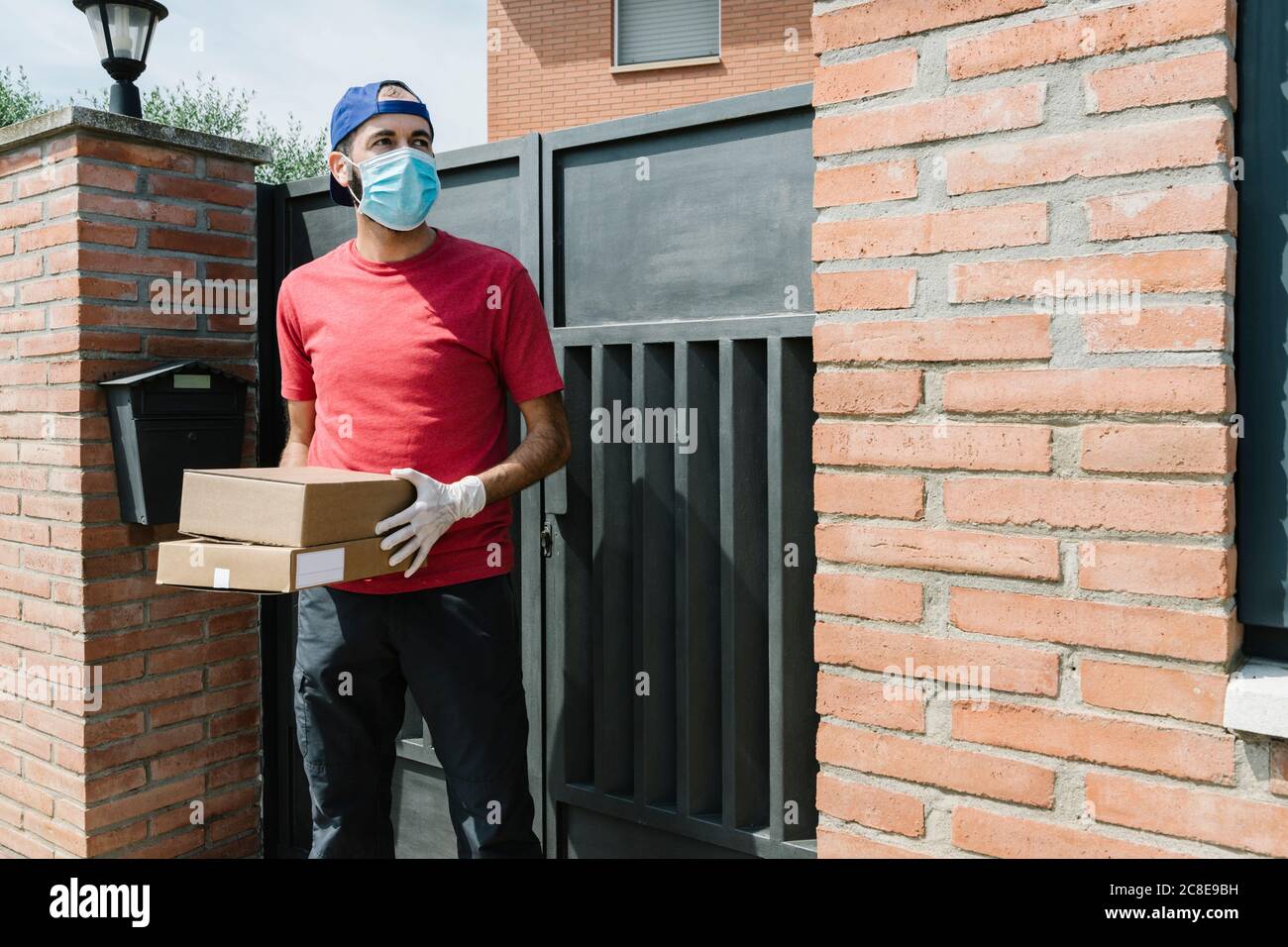 Mid adult male delivery person looking away while holding package and standing against house gate Stock Photo
