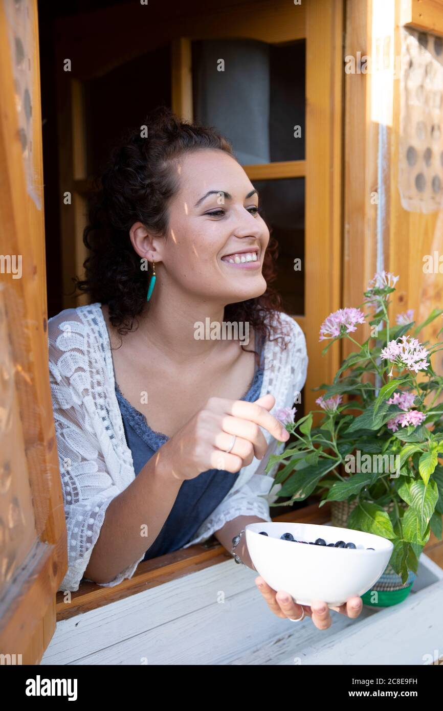 Happy woman holding blueberries in bowl while peeking through window of cottage Stock Photo