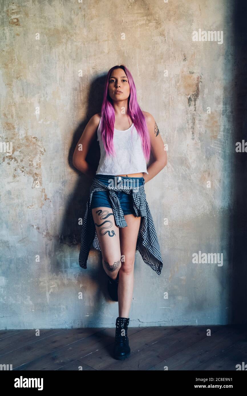 Portrait of a stylish young woman with pink hair leaning against a wall ...