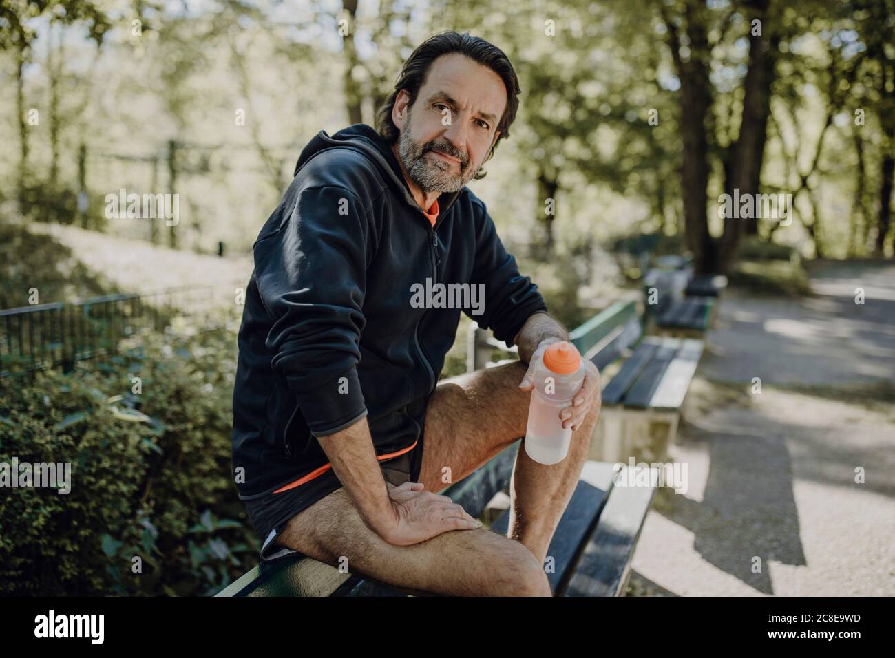 Confident mature man holding water bottle while sitting on bench in park Stock Photo