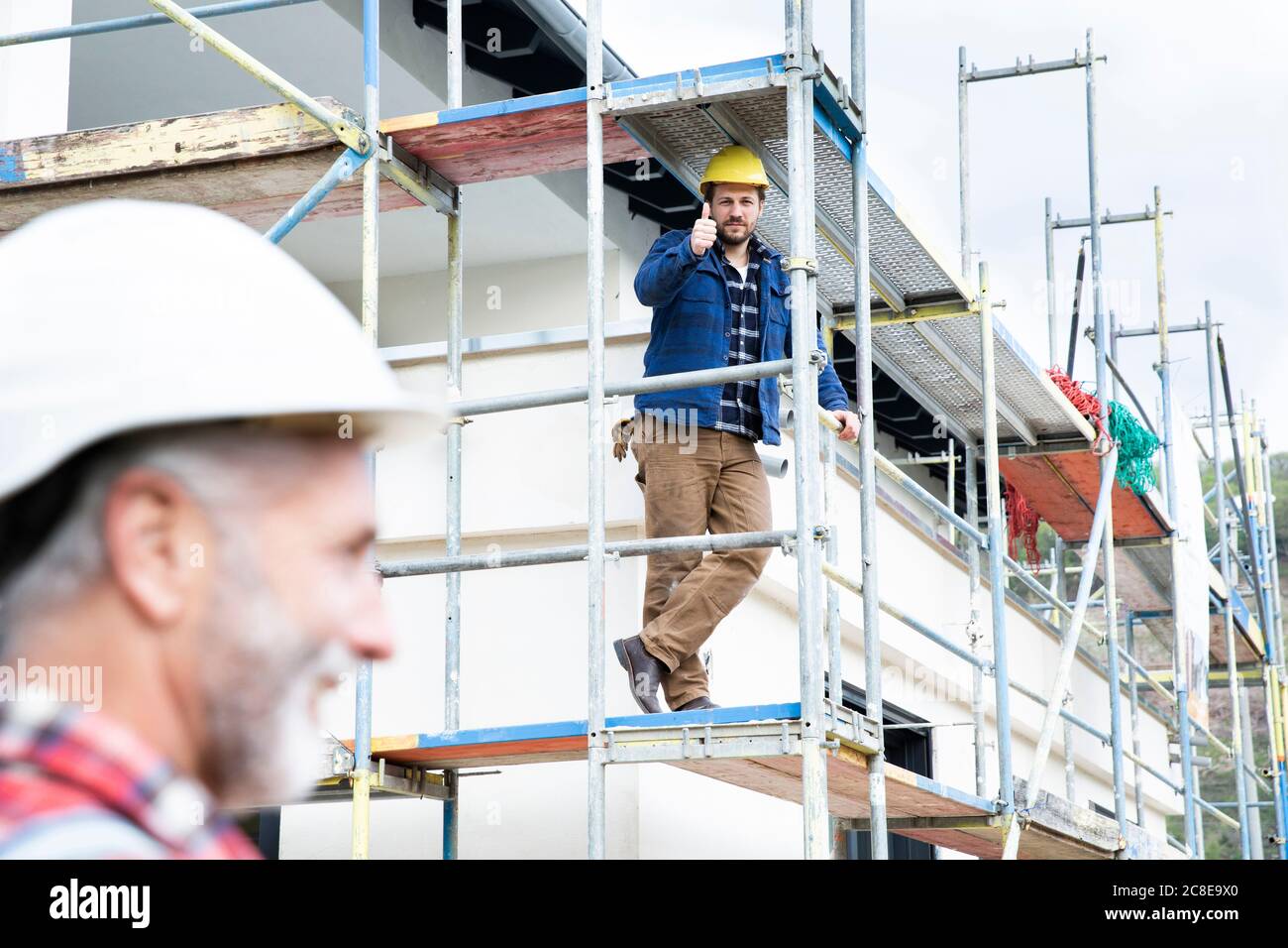 Construction worker showing thumbs up to coworker while sitting on scaffold at site Stock Photo