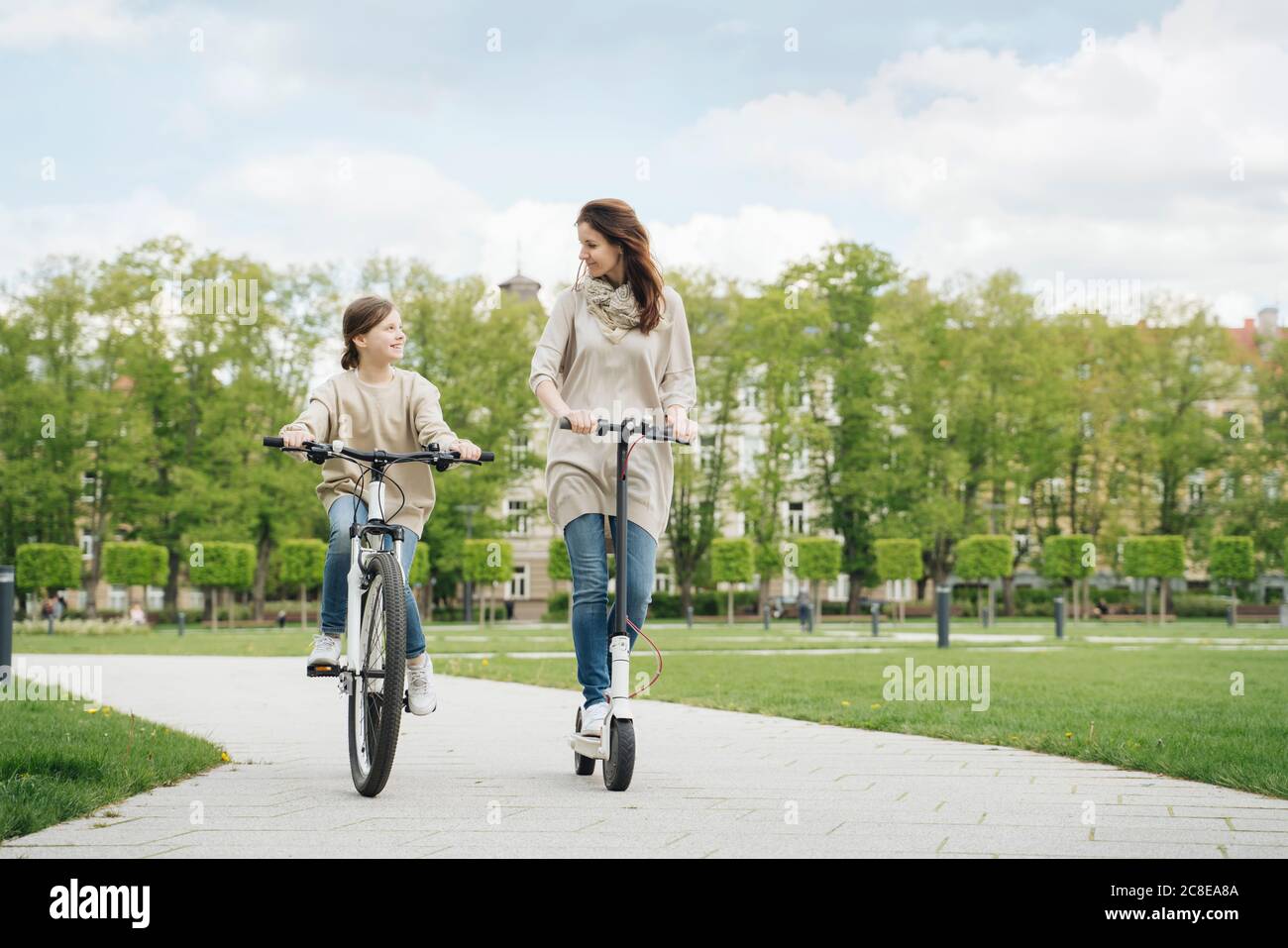 Girl cycling while mother riding electric scooter in city park Stock Photo