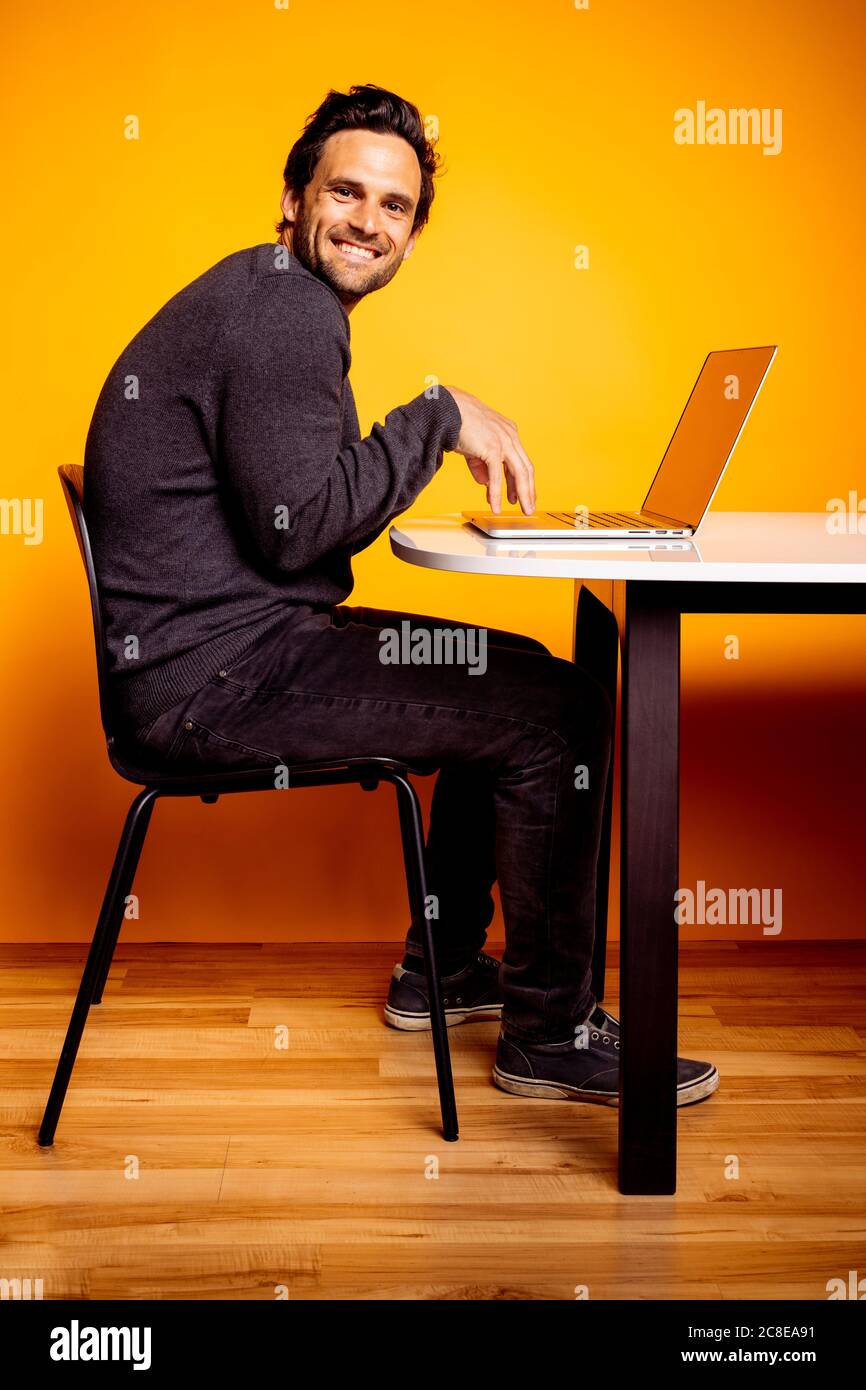 Smiling handsome man using laptop while sitting at table against yellow background Stock Photo