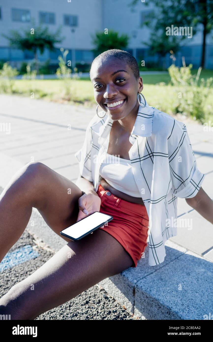 Smiling young woman with shaved head using mobile phone while sitting on footpath in city Stock Photo
