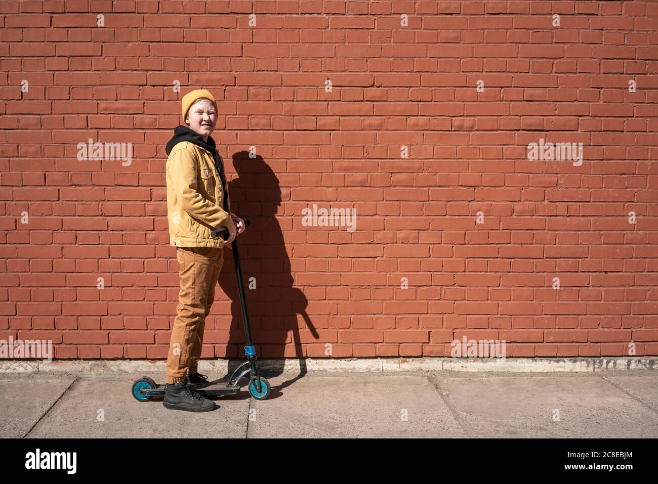 Boy winking while standing with push scooter on sidewalk against brick wall Stock Photo