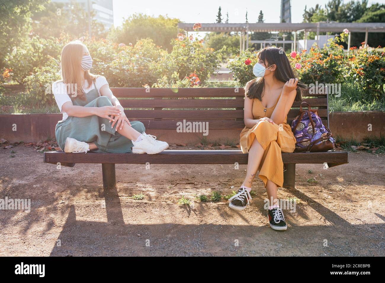 Two friends wearing protective masks sitting on bench during Corona crisis and keeping distance Stock Photo