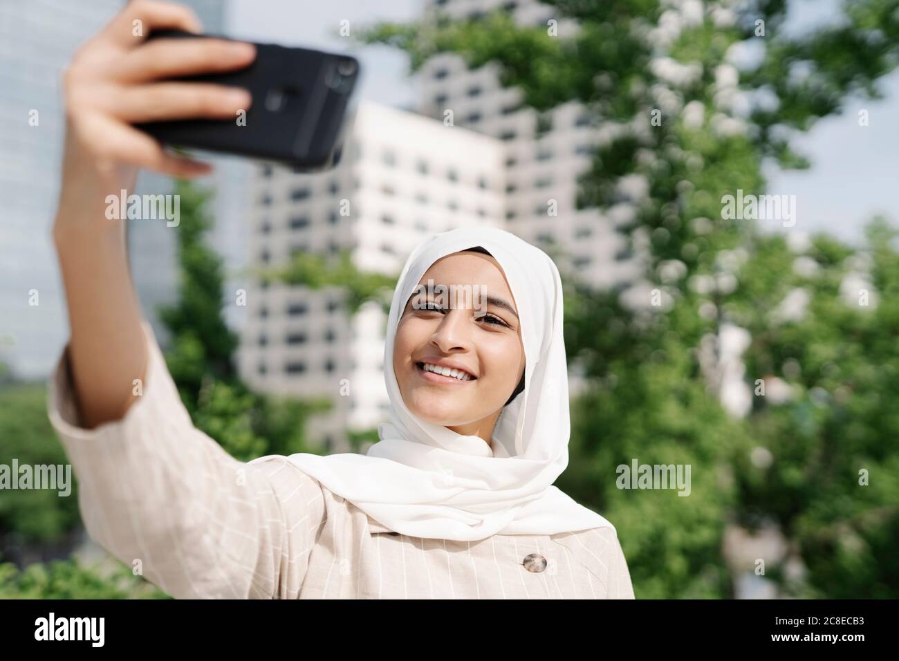 Muslim teenage girl smiling while taking selfie through smart phone in city Stock Photo