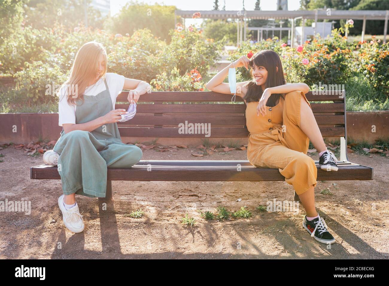 Two friends with protective masks sitting on bench during Corona crisis keeping distance Stock Photo