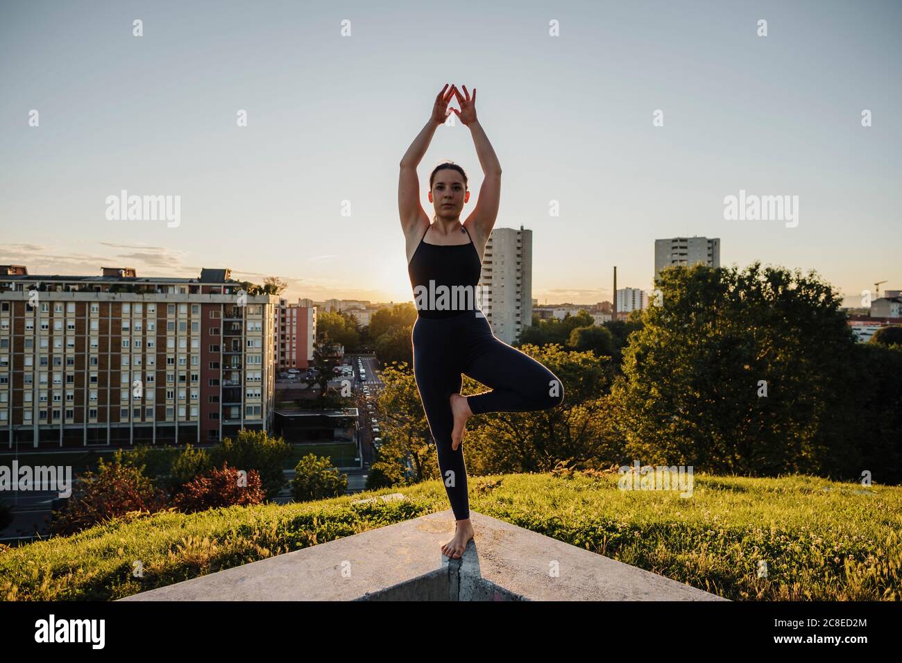 young woman practicing yoga in tree pose on retaining wall in city during sunset 2C8ED2M
