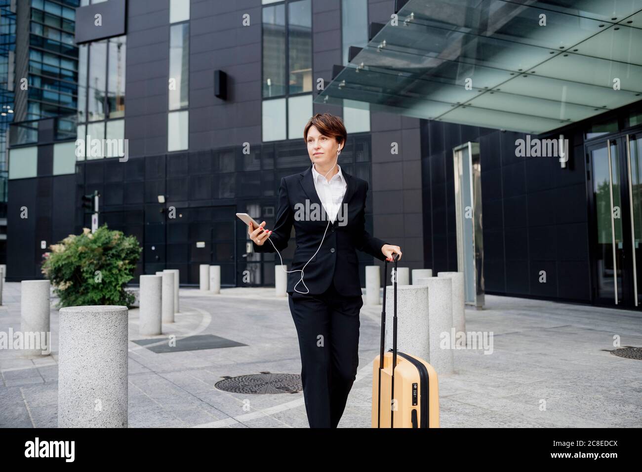 Female professional listening music while walking with suitcase against modern building Stock Photo