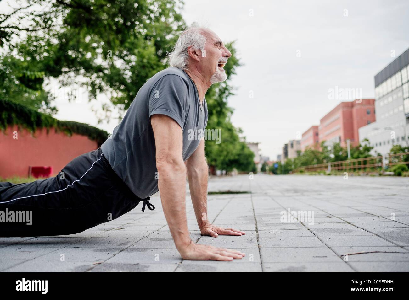 Senior man with mouth open doing push-ups on footpath in city Stock Photo