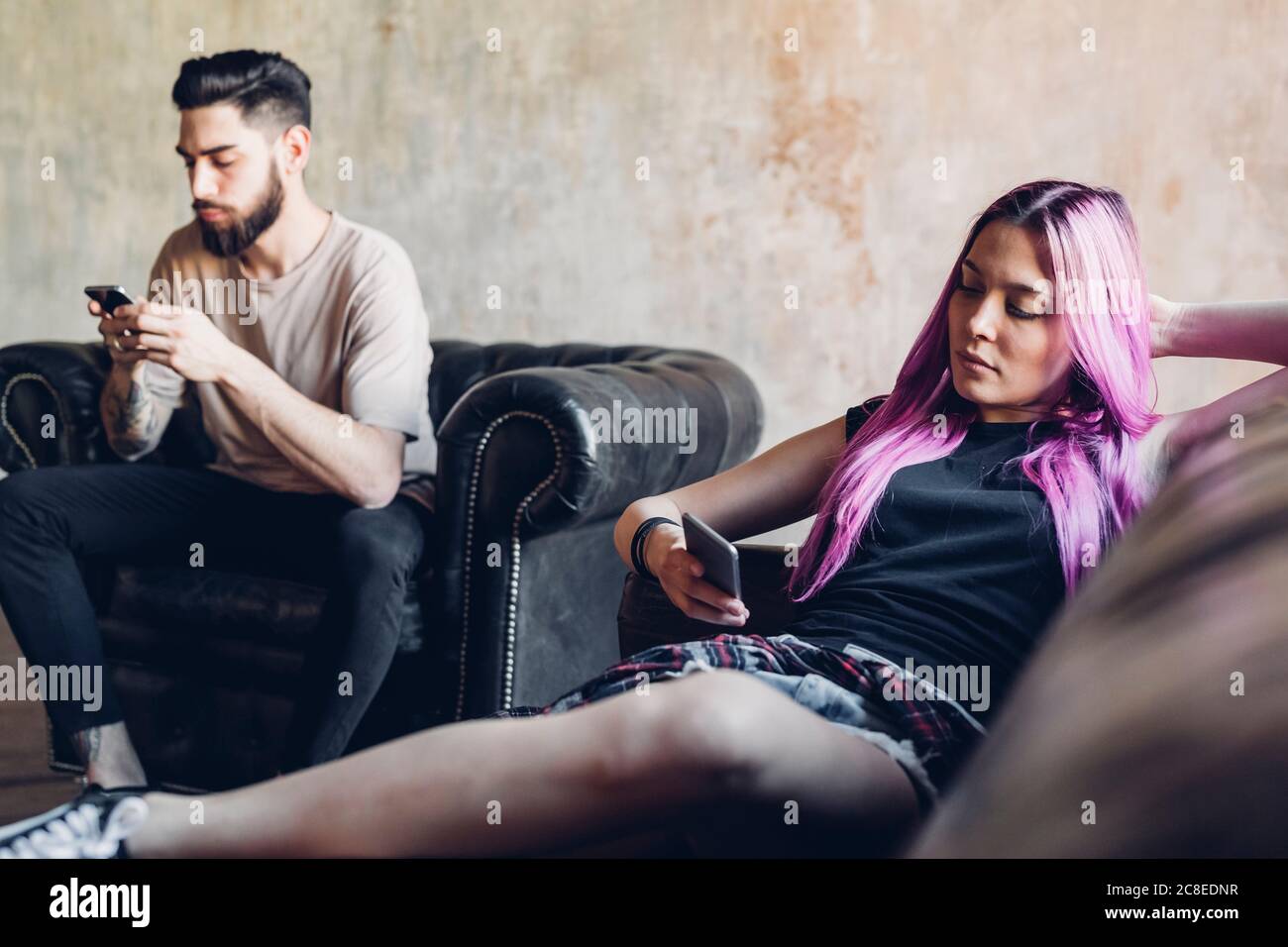Young man and woman sitting on sofa and armchair in a loft using smartphones Stock Photo