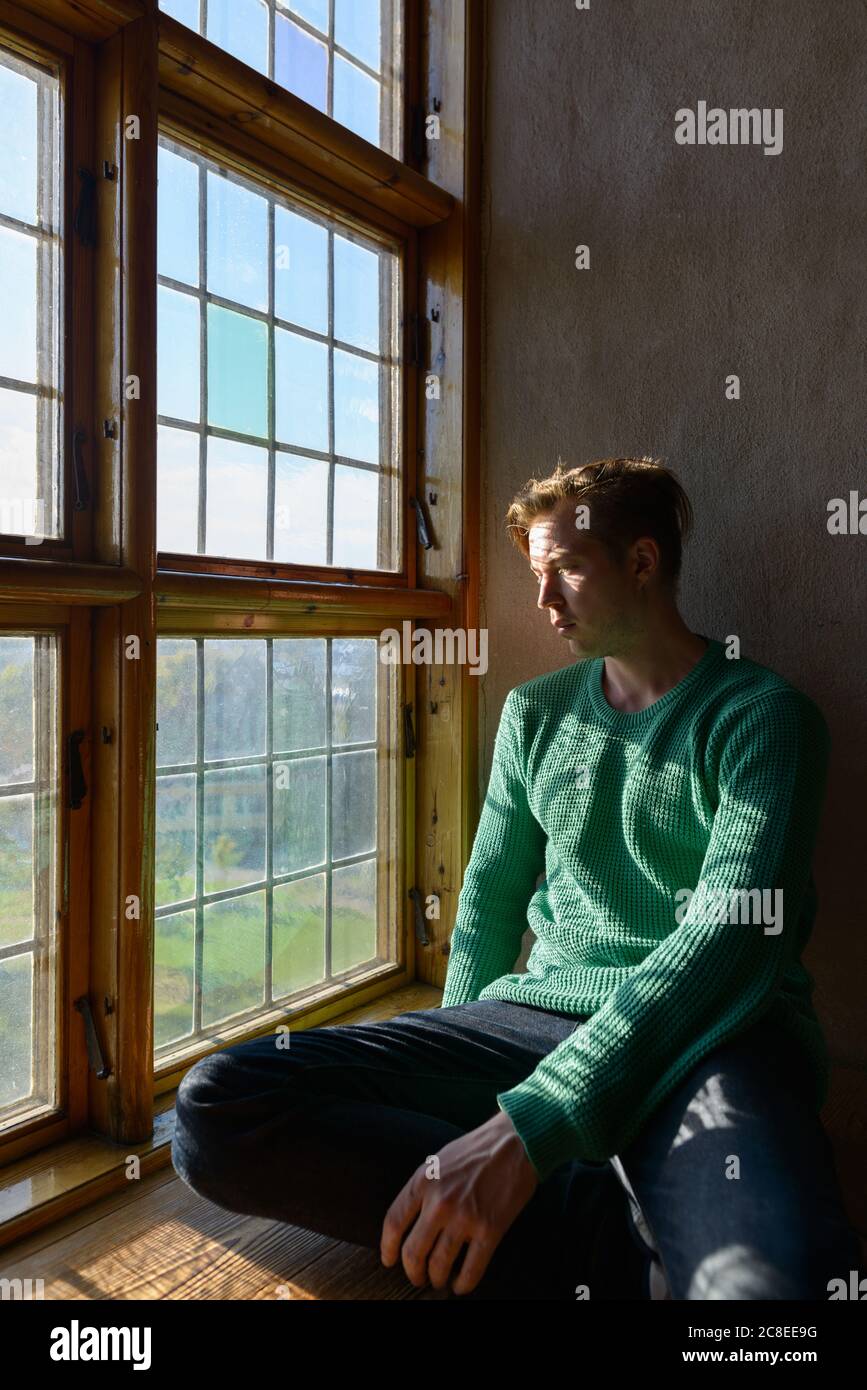 Young handsome man sitting in front of closed wooden window with sunlight streaming in Stock Photo