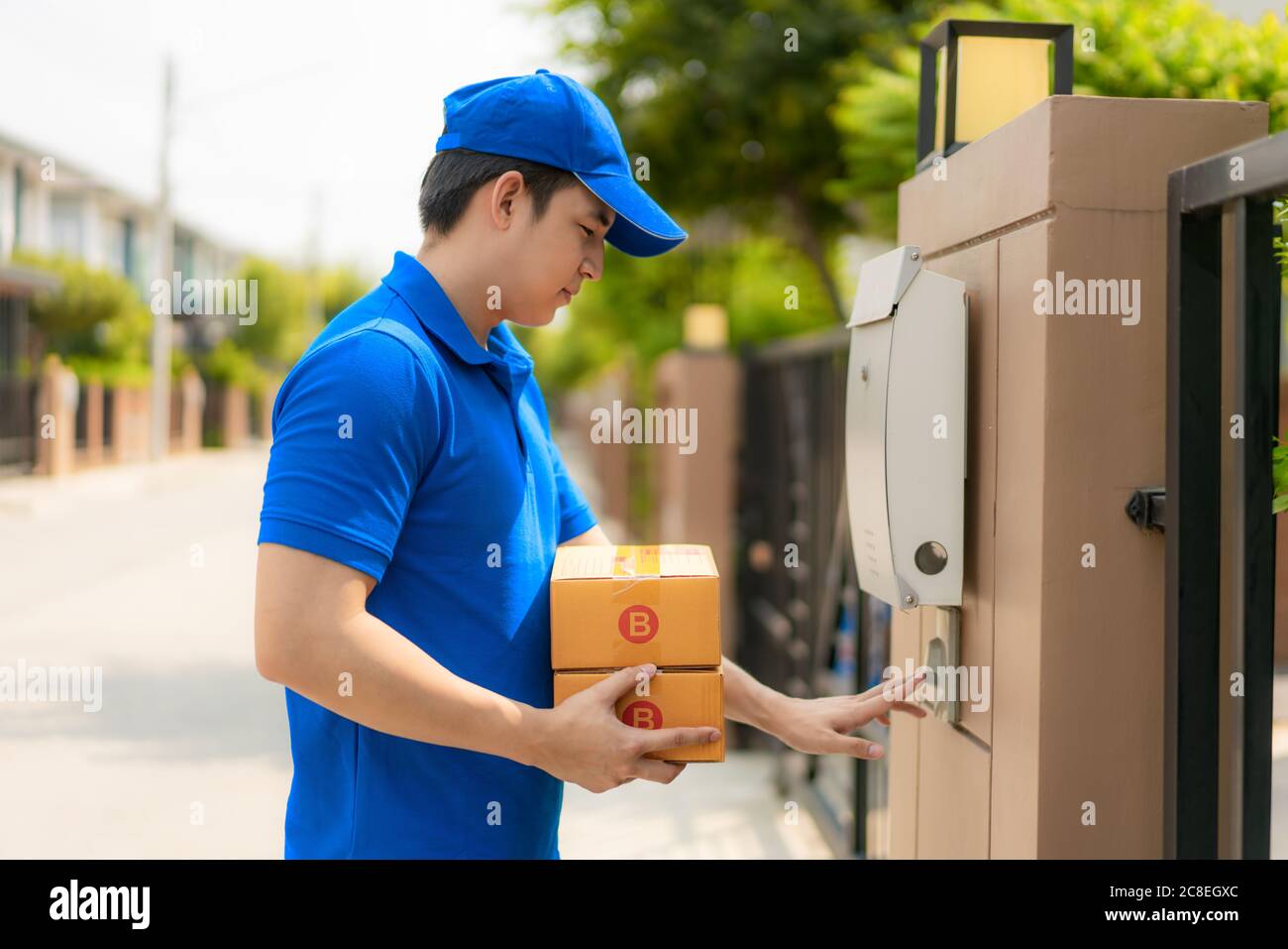 manAsian delivery young man in blue uniform with parcel box smile and  ringing customers door bell in front house village with copy space. Advertising Stock Photo