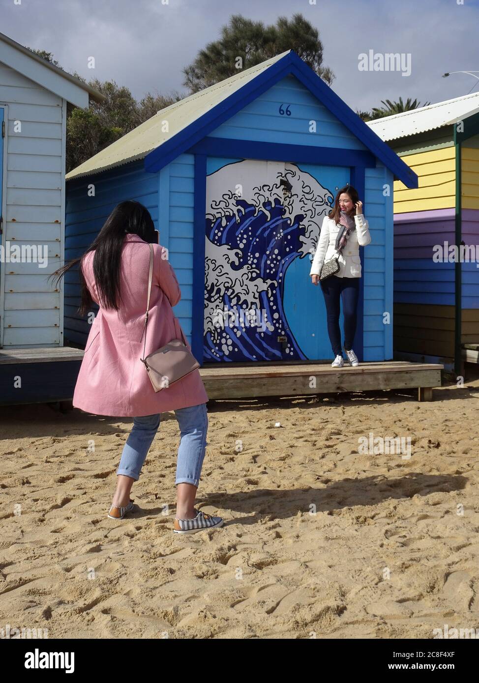 Female tourists, unsuitably dressed for the beach, use bathing boxes on the Dendy Street Beach, Melbourne, Australia, as a backdrop for their 'selfies Stock Photo