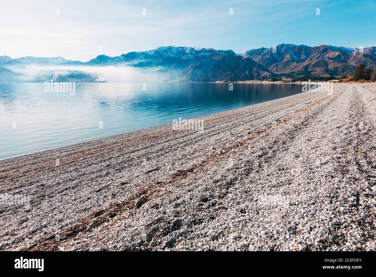 A calm, clear sunny winter day on Scotts Beach, Lake Hawea in the South Island of New Zealand. A farm burn off casts a layer of smoke across the bay Stock Photo
