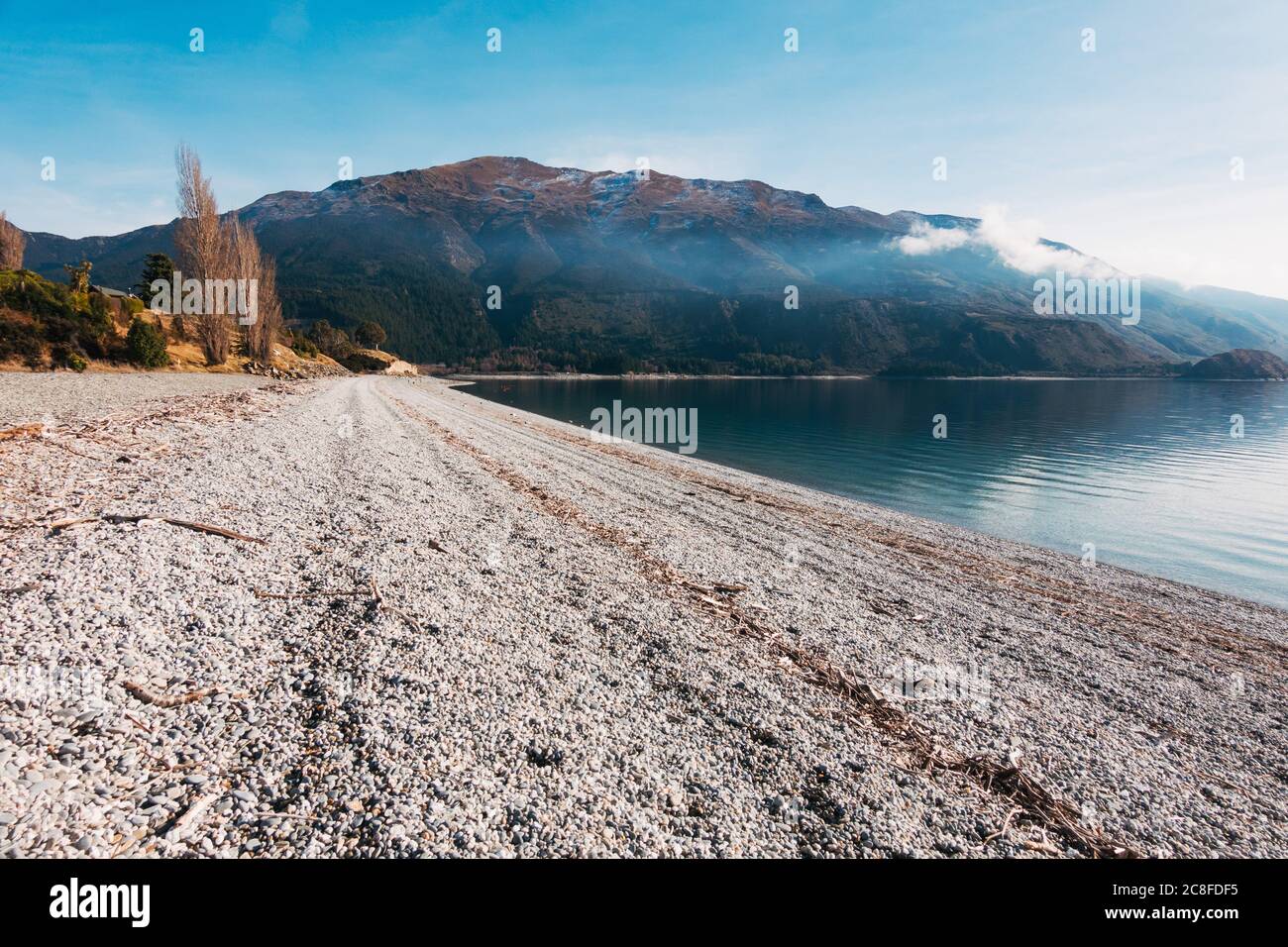 A calm, clear sunny winter day on Scotts Beach, Lake Hawea in the South Island of New Zealand. A farm burn off casts a layer of smoke across the bay Stock Photo