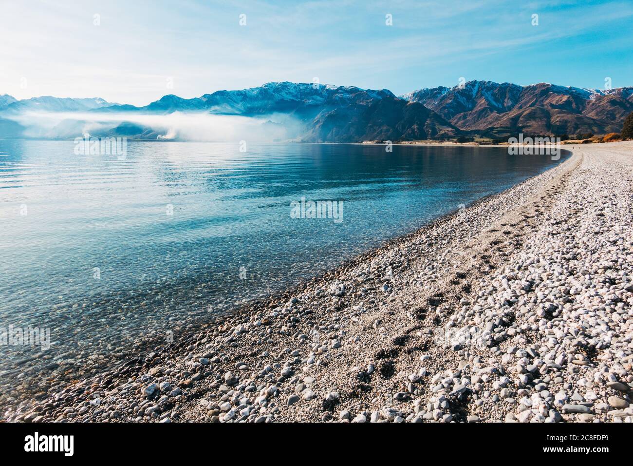 A calm, clear sunny winter day at Lake Hawea in the South Island of New Zealand. A farm burn off casts a layer of smoke across the bay Stock Photo