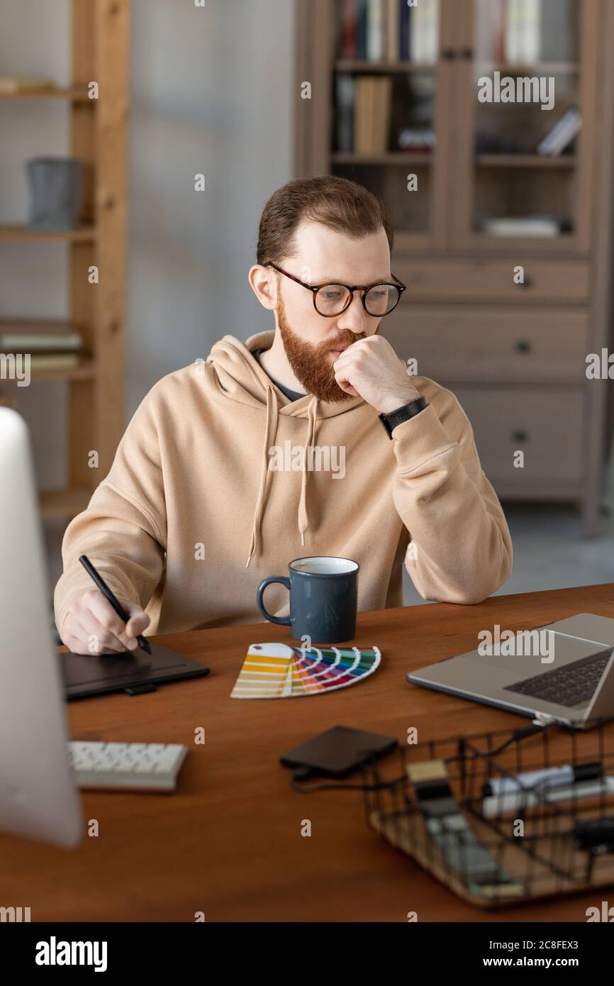 Thoughtful young designer in hoodie sitting at wooden table and ...