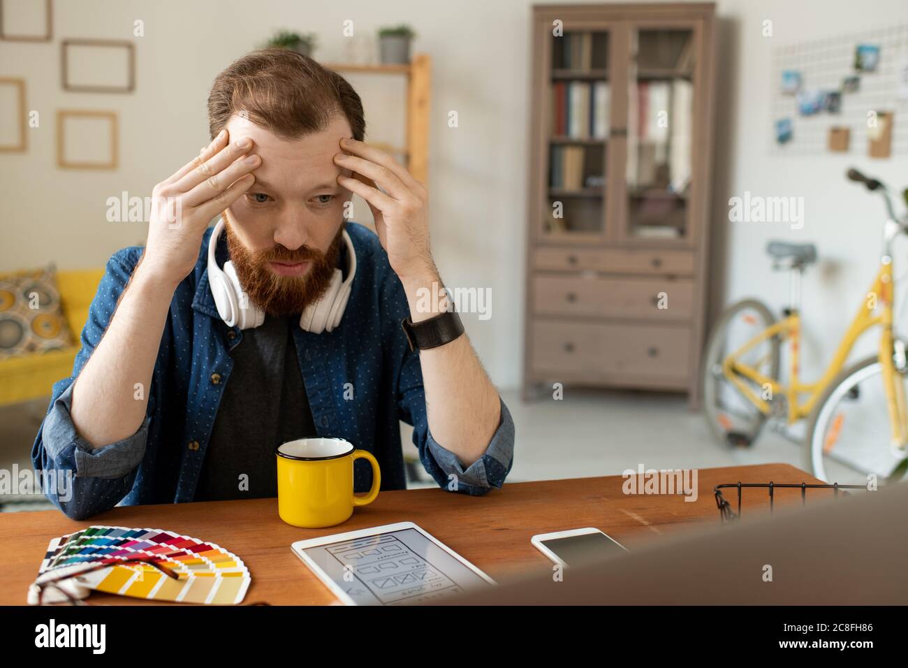 Puzzled young UI designer with beard sitting at table and holding head in hands while thinking of interface layout Stock Photo