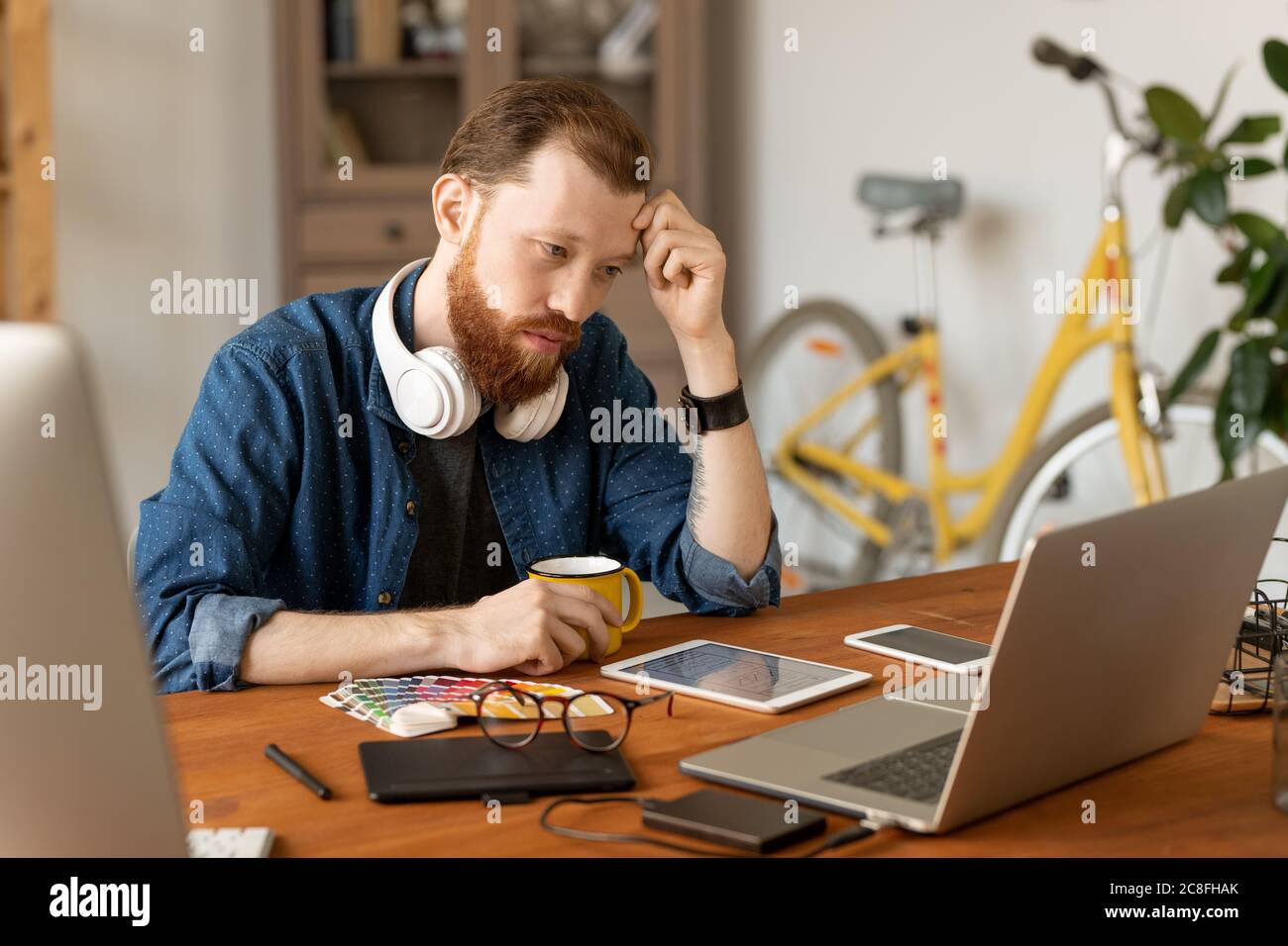 Serious young bearded man focused on design concept sitting at desk and looking at sketch on laptop screen Stock Photo