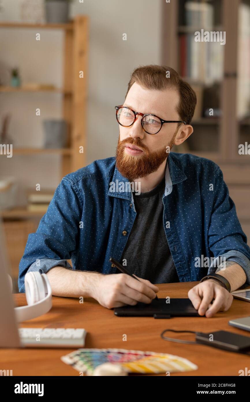 Hipster bearded man in eyeglasses sitting at table and using digitizer tablet while thinking of visual solution Stock Photo