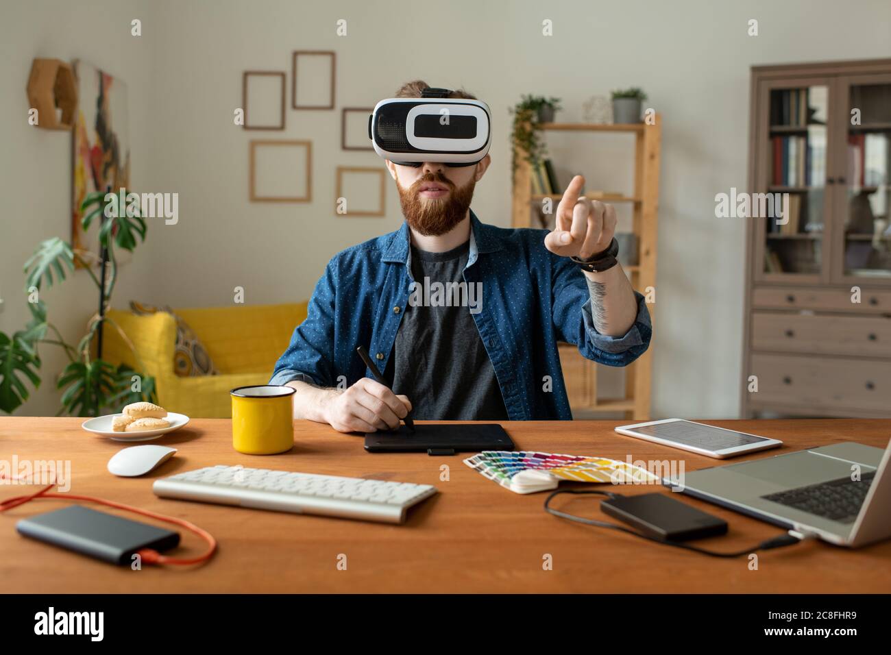 Serious bearded designer sitting at table and drawing on digitizer while using virtual reality simulator at home Stock Photo