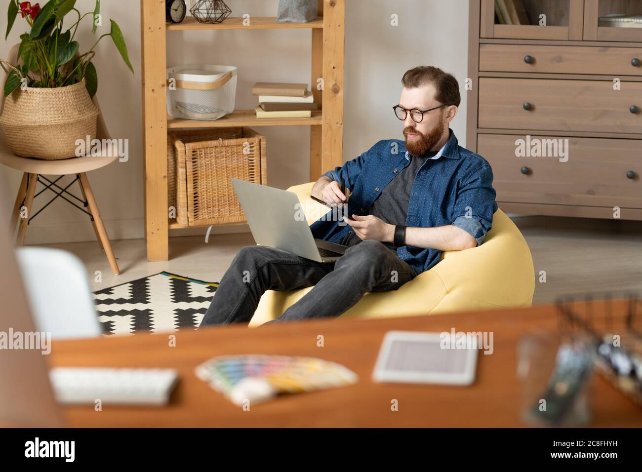 Confident young layout artist with beard sitting in yellow bean bag and