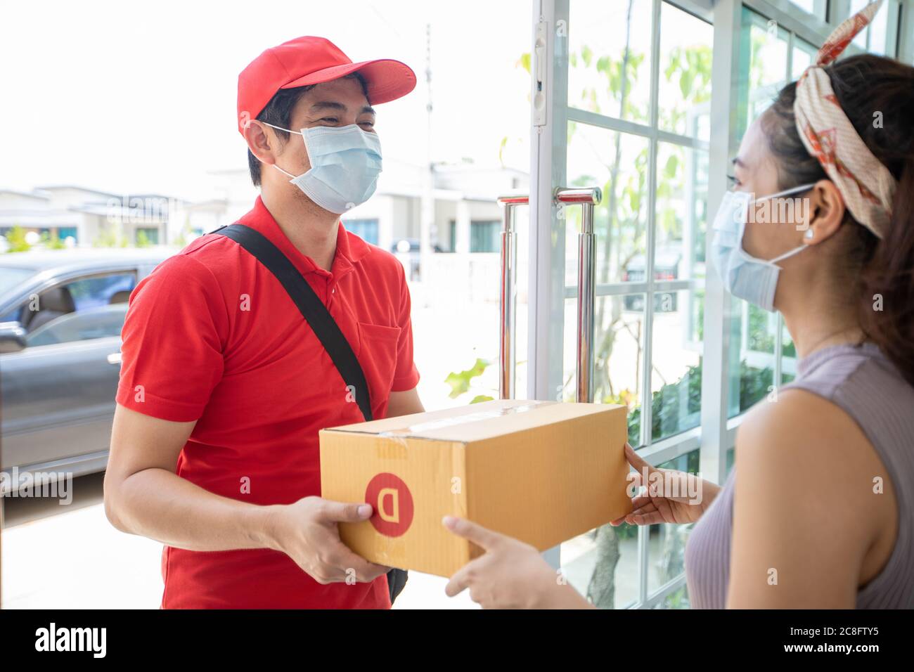 Asian delivery servicemen wearing a red uniform with a red cap and face mask handling cardboard boxes to give to the female customer in front of the h Stock Photo