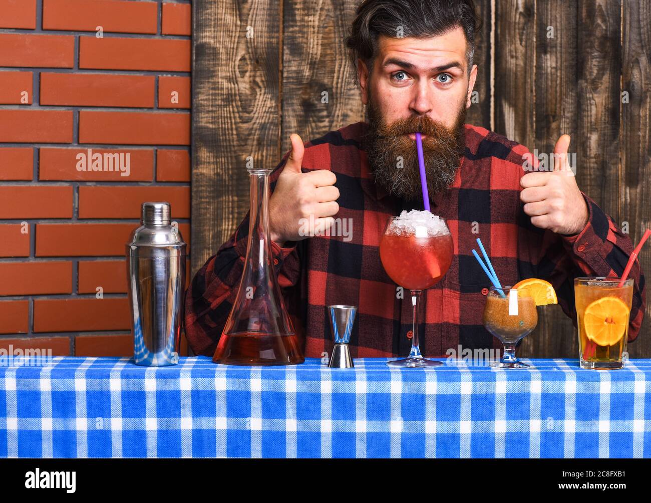 Barman with beard and cheerful face drinks out of glass with drinking straw cocktail. Man in checkered shirt on wooden background. Barman and cocktails concept. Hipster enjoy drink and thumbs up. Stock Photo