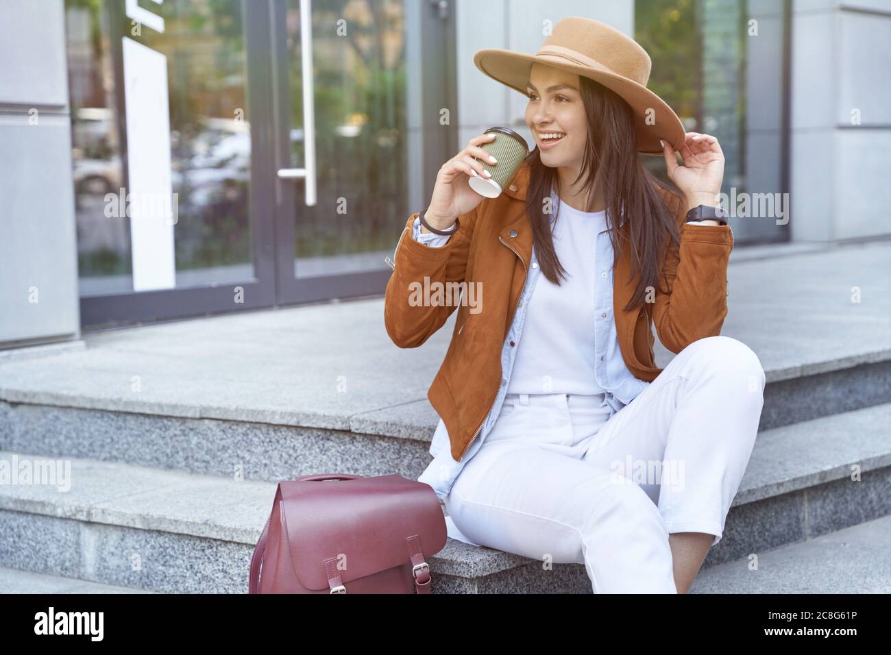 Enjoying vacation. Young and beautiful happy female traveler or tourist wearing hat and backpack drinking coffee and smiling while resting on city stairs. Travel and tourism concept Stock Photo
